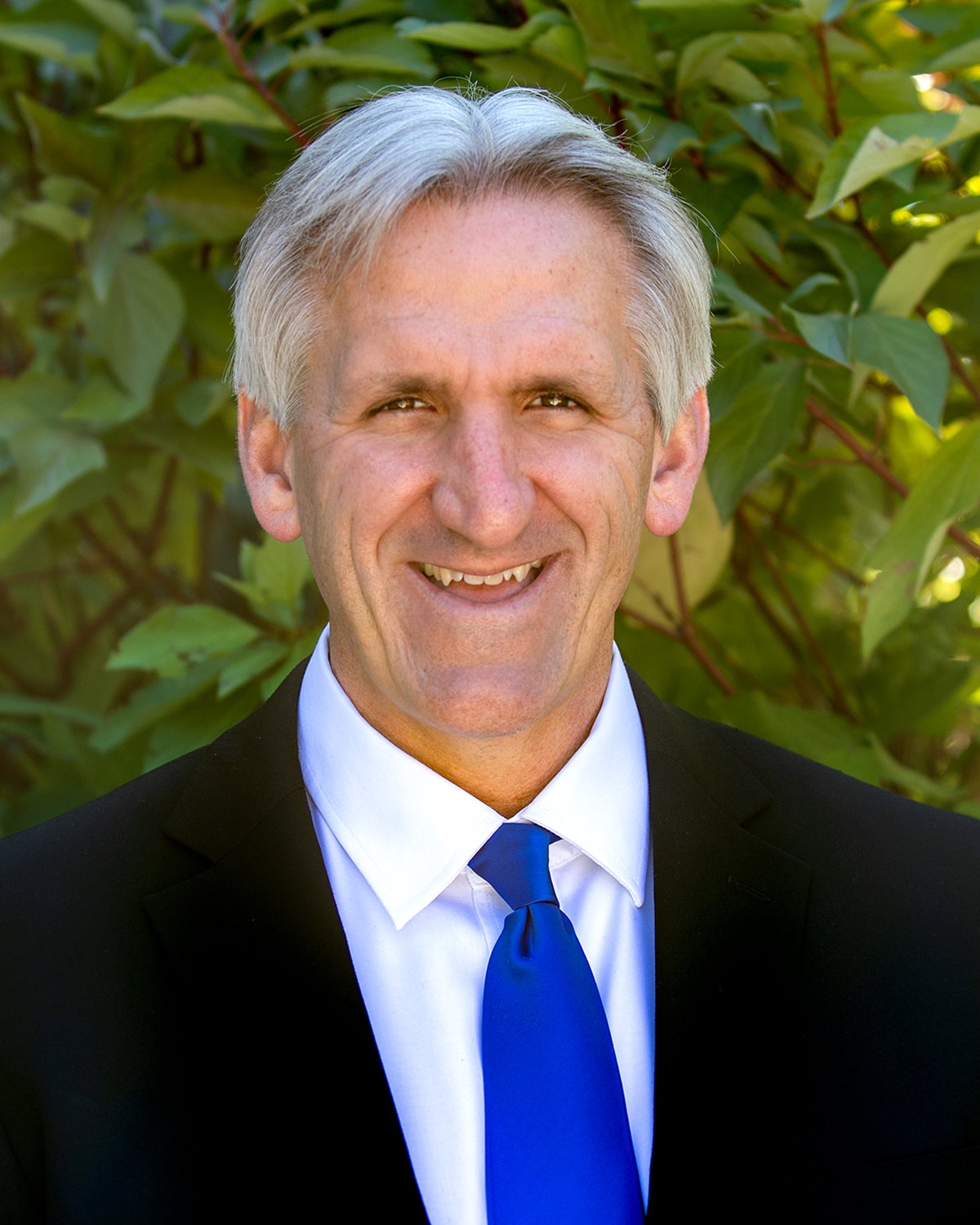 Provost John Buckwalter, wearing a suit with a blue tie, smiling, with leaves in the background