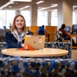 A student seated at a table smiling while using their laptop.
