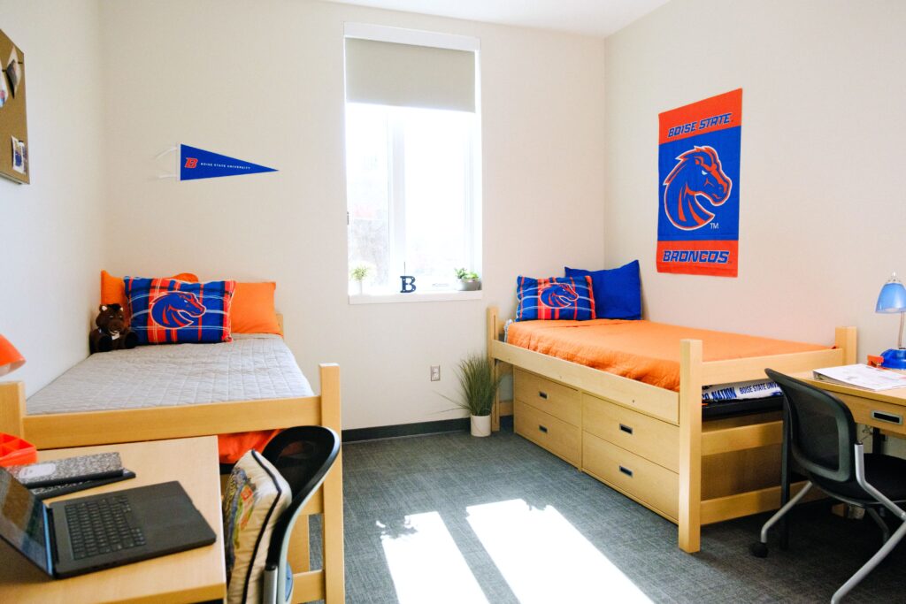 A double dorm room showing two beds on either side of the room with Boise State branded pillows, and a poster that reads 'Boise State Broncos' on one wall.