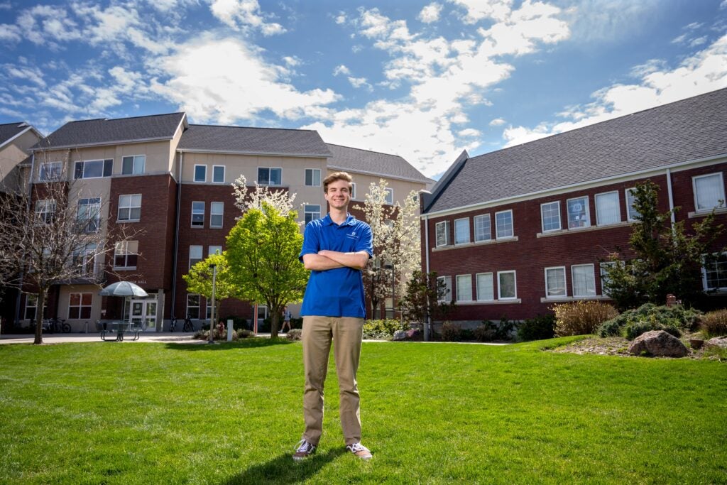A student standing outside on grass, with trees and two residence hall buildings behind them.