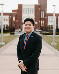 Isaac Celedon standing in front of a brick building smiling with hands clasped in front of him