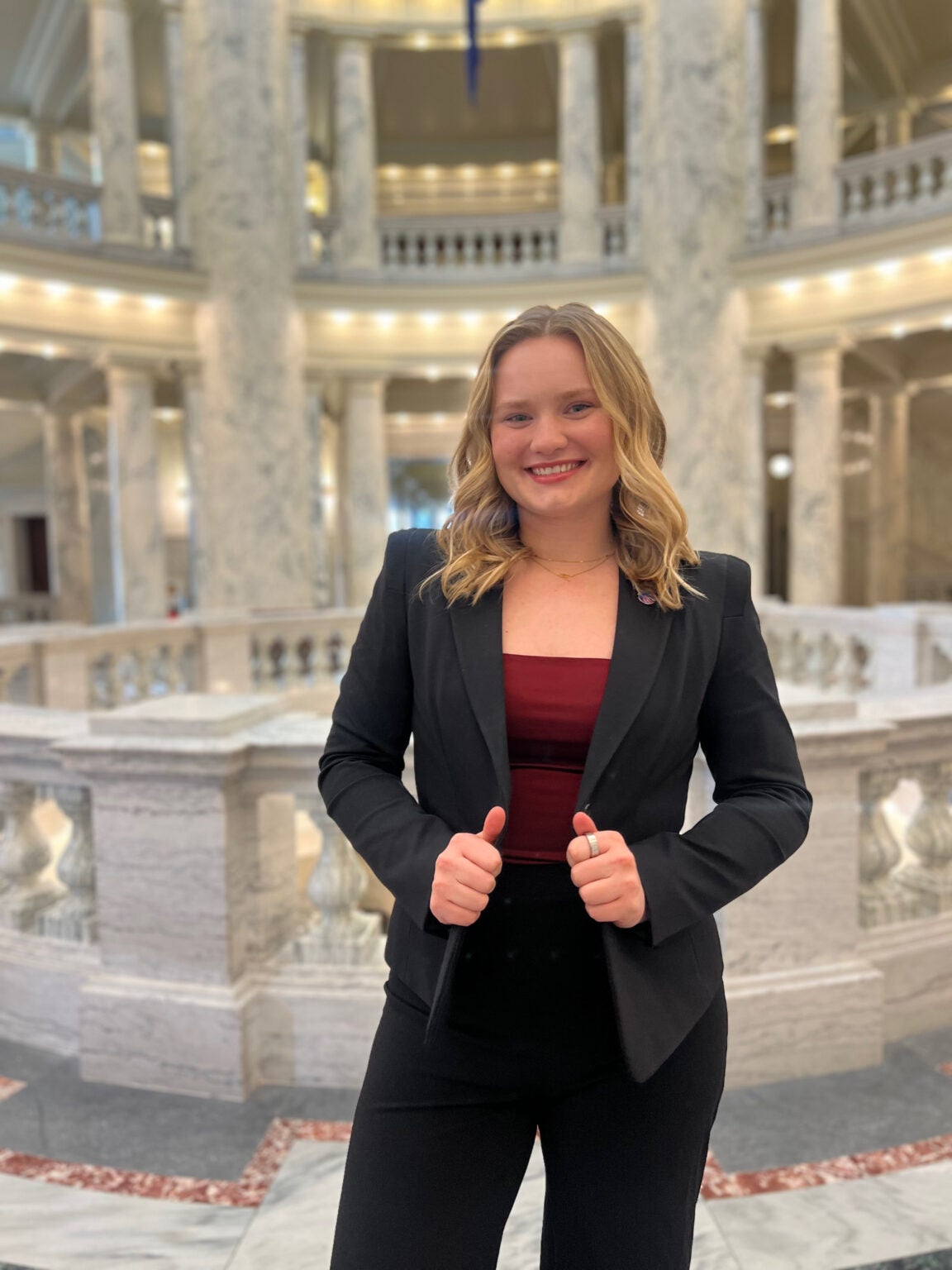 Maggie Tell wearing a crimson blouse and grey blazer, smiling and standing in the rotunda of the Idaho State capitol