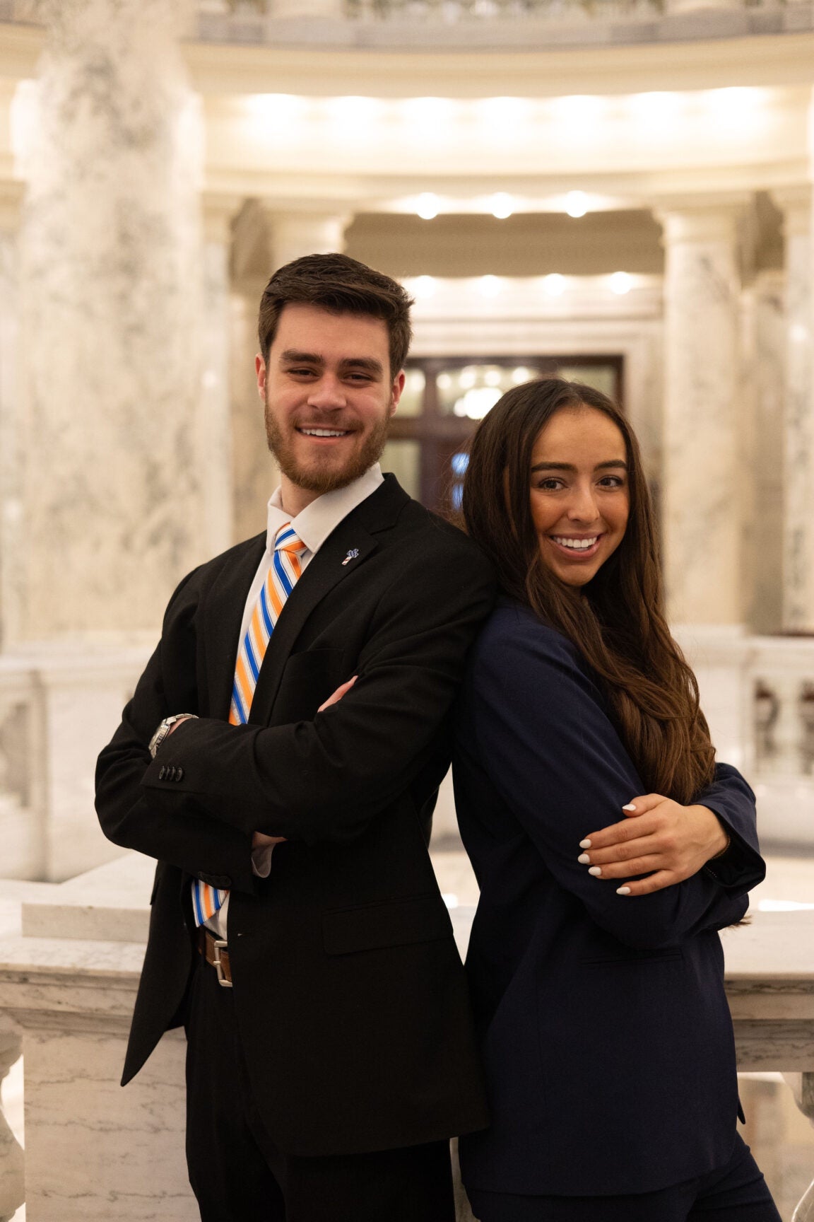 Austin Rose and Abby Berndt in the Idaho Capitol Rotunda, standing back to back with arms folded, smiling