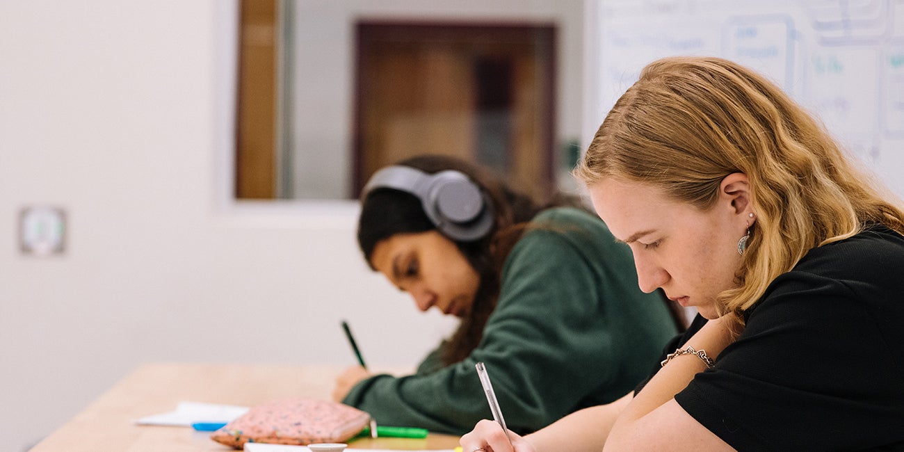 two students working intently at a desk, writing on papers, one is wearing headphones