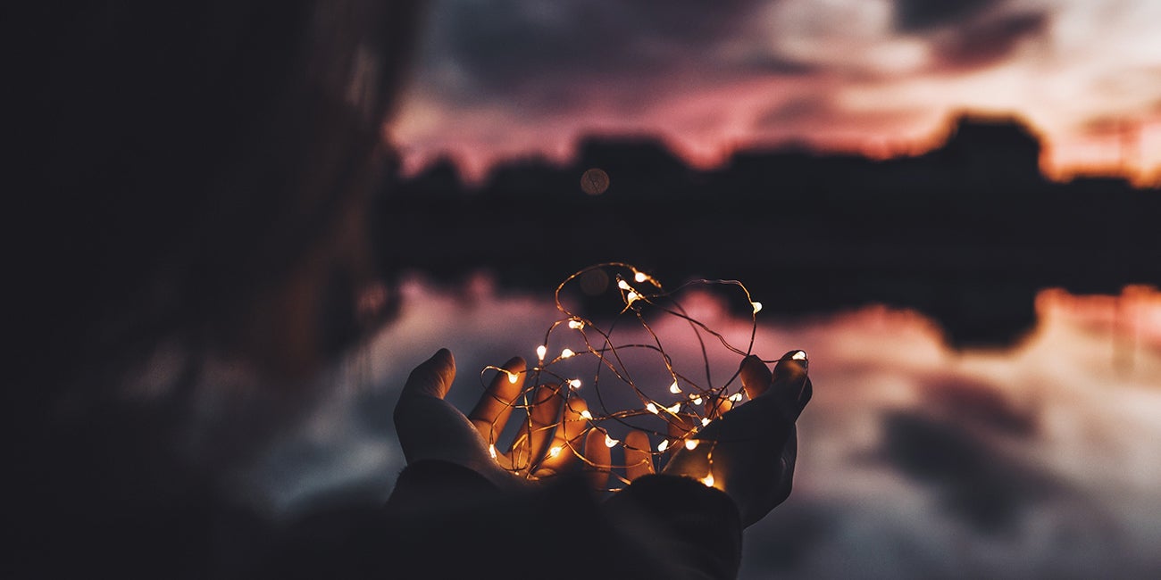 Cupped hands holding glowing fairy lights against a blurred twilight water scene.