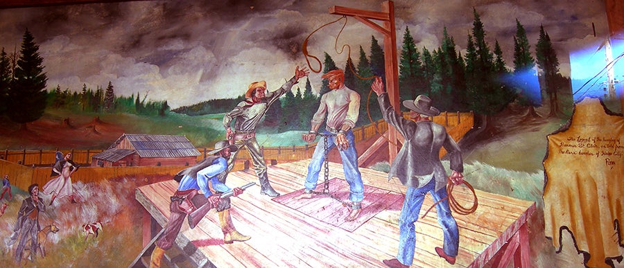 Man stands chained under a hangman's noose, one man points a gun while two others set up the rope