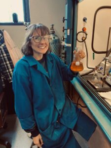 Photo of Maddelyn Glassey in a lab, wearing a blue lab coat and holding a beaker filled with yellow liquid
