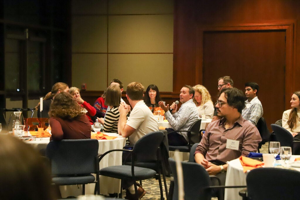 A lively discussion is taking place in a well-lit conference room where attendees are seated around banquet tables; one man actively speaks into a microphone, gesturing with his other hand, while the rest of the group listens, some looking at the speaker and others conversing among themselves.