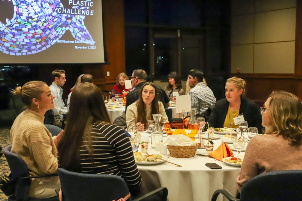 A group of professionals is engaged in a discussion at a roundtable event, with a presentation screen in the background titled "PLASTIC CHALLENGE" from the Fall 2023 Business Ethics Roundtable, focusing on sustainable practices for McDonald's restaurants.
