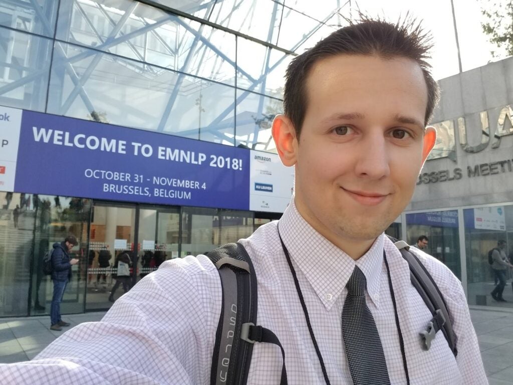 Selfie of Dan Kondratyuk, with a sign behind him saying "Welcome to EMNLP 2018!"