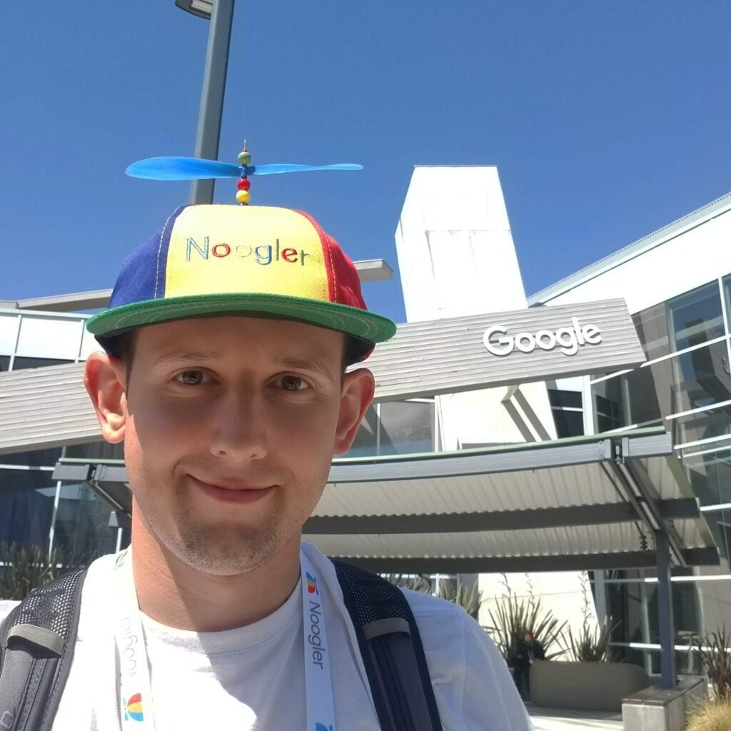 Selfie of Dan Kondratyuk, standing left of a Google building while wearing a funny hat that says 'Noogler'.