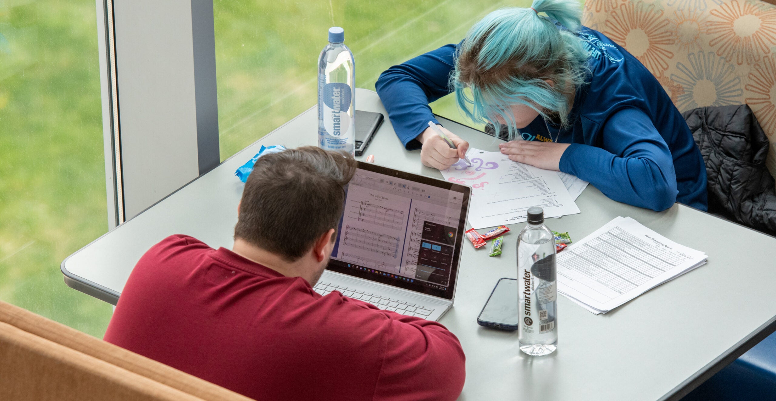 Students lean over table writing on paper and working on laptops