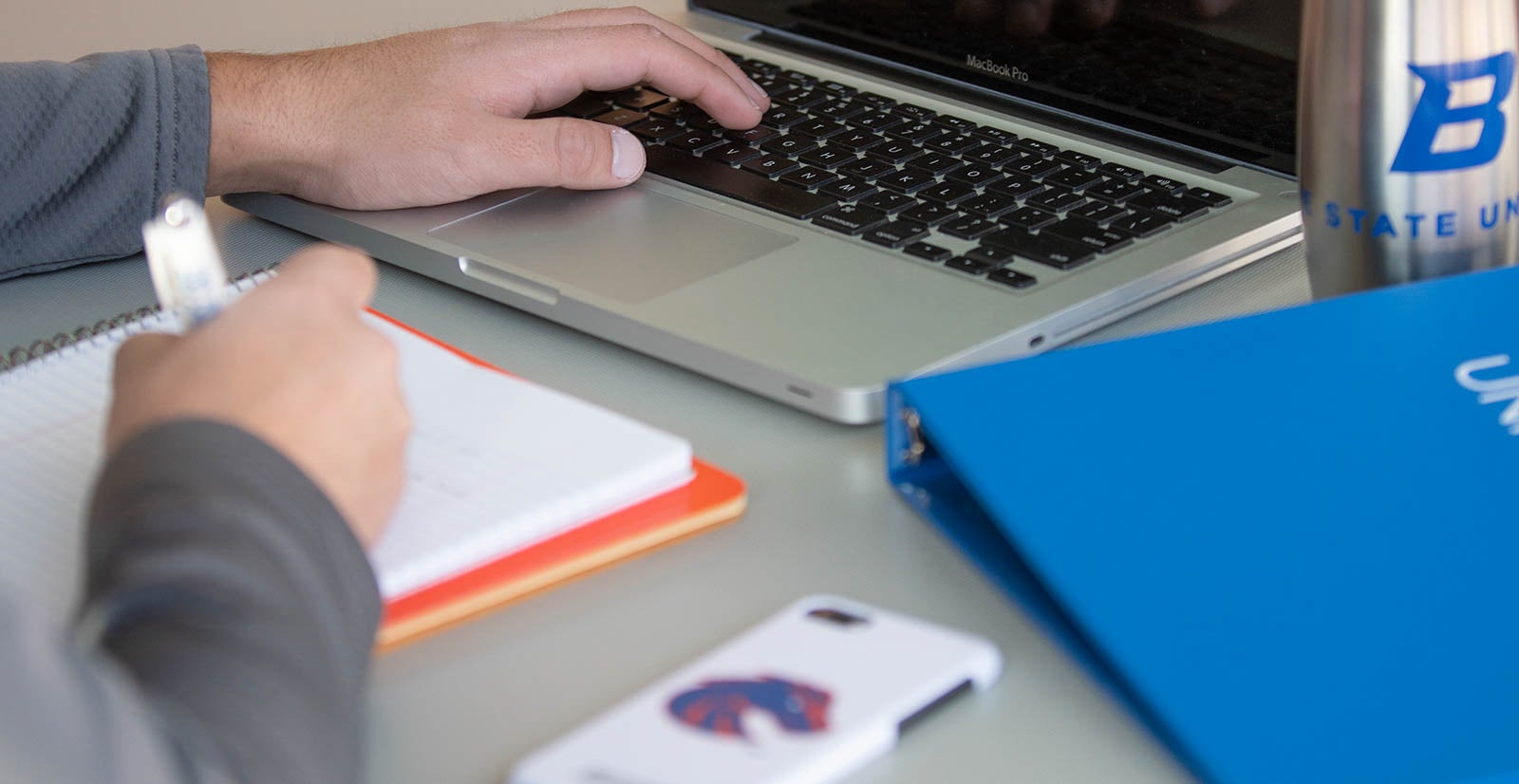 Student types on laptop while writing on notebook