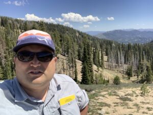 A man in a hat and sunglasses in front of a forested mountain hillslope with blue skies and white cumulus clouds in the background.