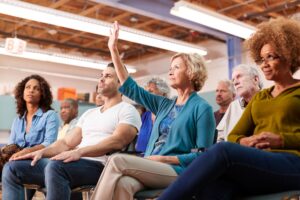 Image of diverse group of people sitting in a meeting, looking engaged. One has a raised hand.