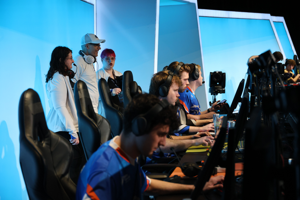 Photo of varsity esports players competing on stage in a large esports arena at CECC 2025. Coaches watch the players from behind on stage.