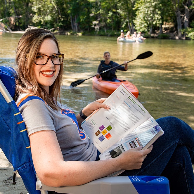 Woman sits by river reading textbook