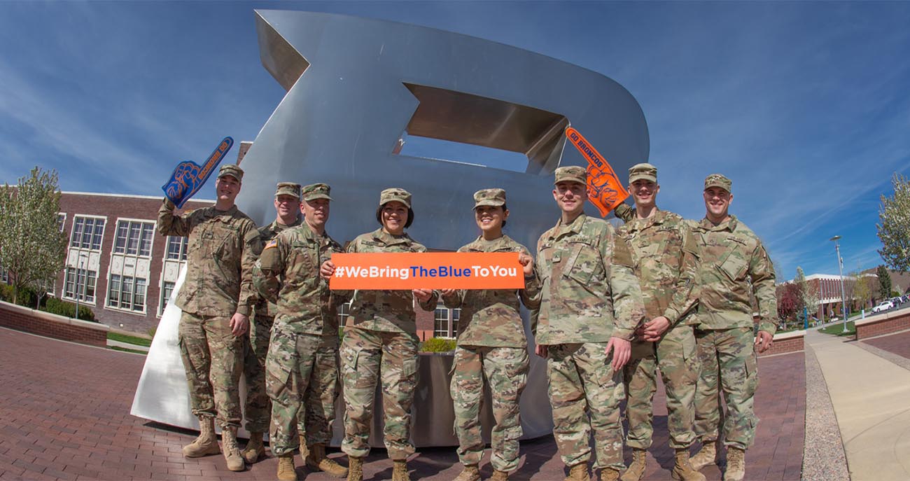 Military students stand in front of the "B" holding a sign saying "We Bring The Blue To You"