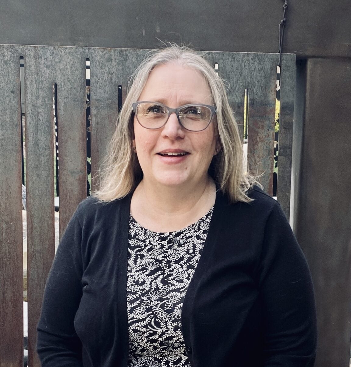 Lana smiles facing the camera. Lana is a white woman with shoulder length blonde hair. She wears glasses and a black and white patterned top. She is standing in front of a fence.