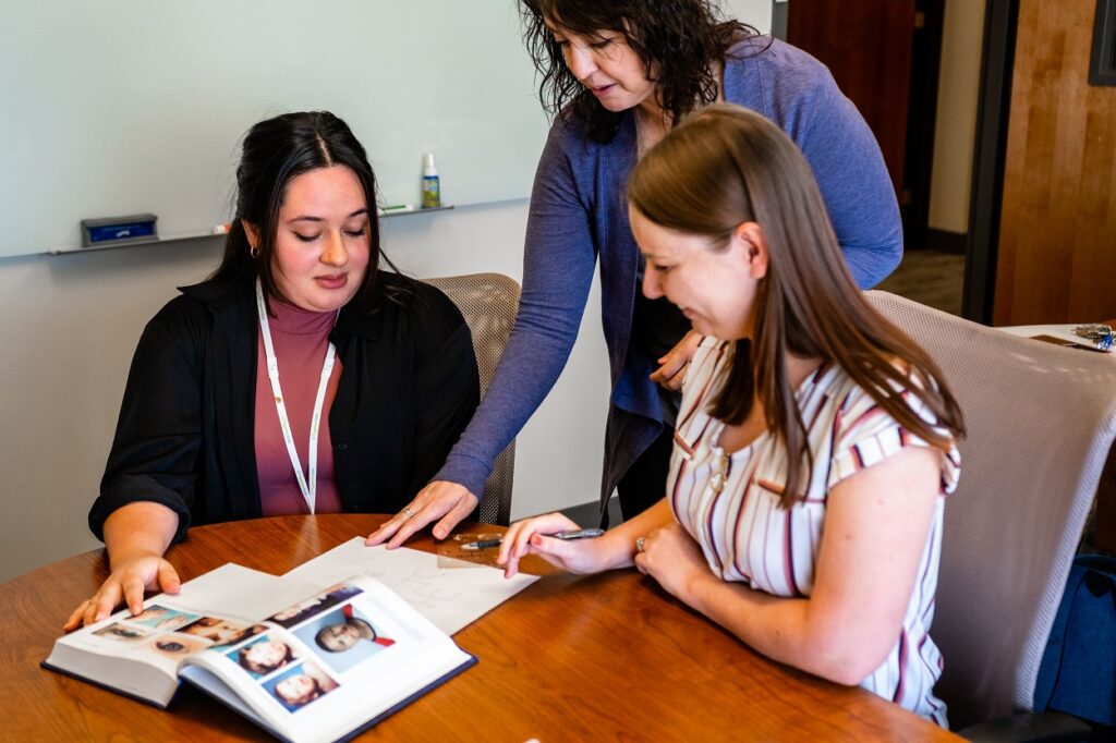 3 women sitting at a table working together examining pictures and text.