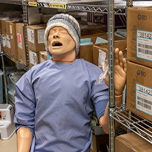 A manikin wearing a knit hat and blue gown sits upright in a storage area