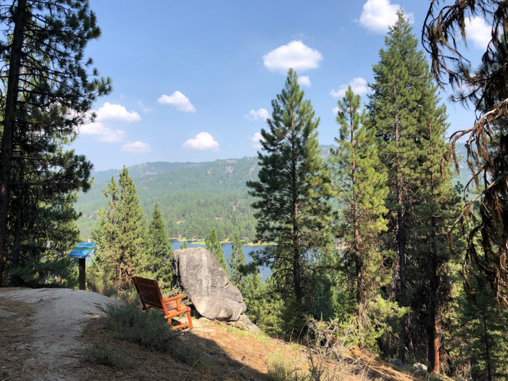 Pine trees border a clearing that overlooks Horsethief Reservoir. In the clearing is a boulder and an informational sign.