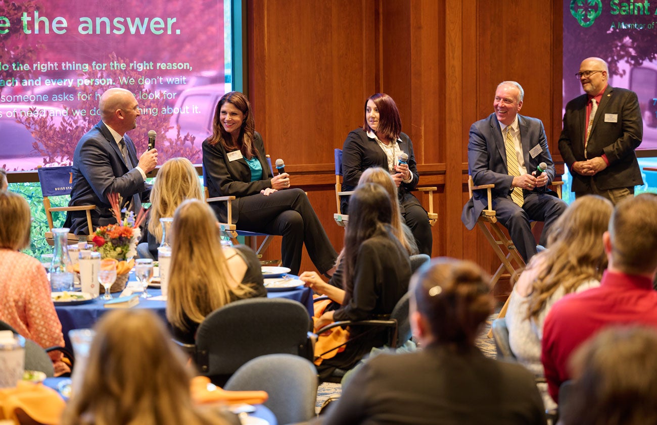 Four people sit in chairs and speak to an audience sitting at tables in front of them.
