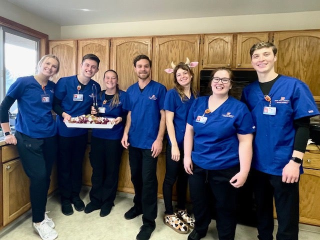 Seven nursing students stand in a kitchen holding a tray of food