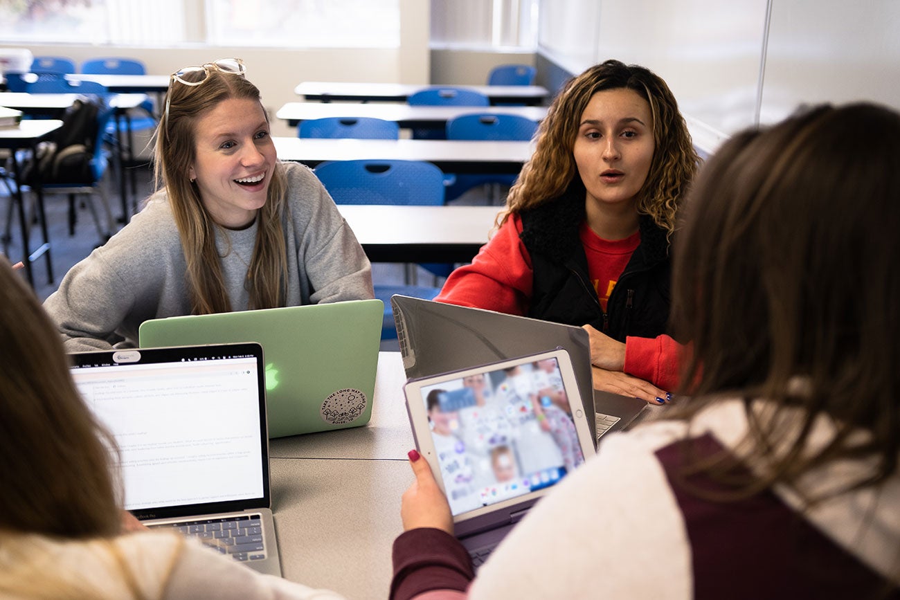 Four students with laptops sit around a table smiling and discussing schoolwork
