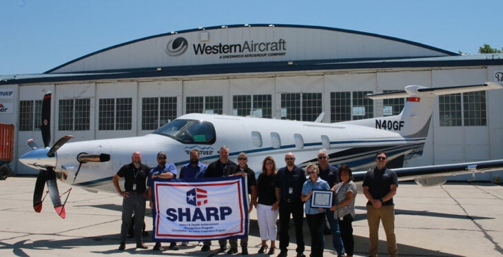 Western Aircraft staff stand with the SHARP flag in front of a plane on the ground in front of a Western Aircraft hanger.