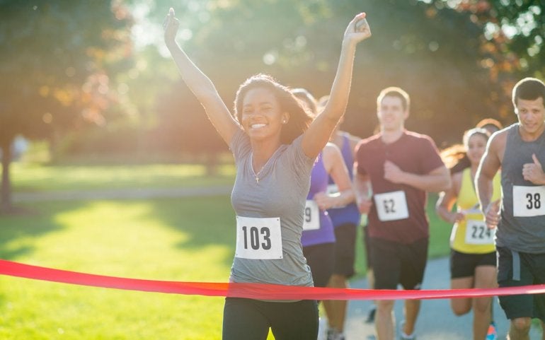 woman running through a red ribbon at finish line of race, her hands in the air and smiling