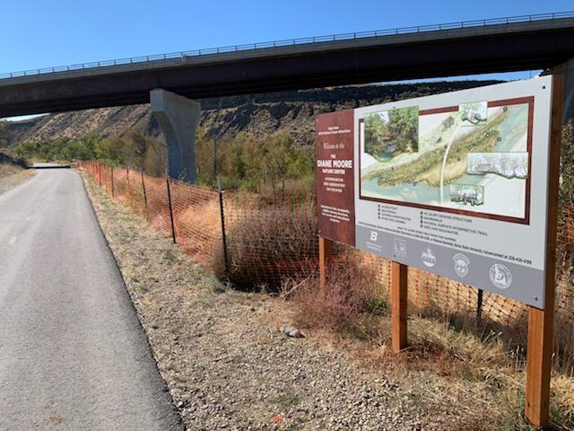 a view of the greenbelt bike path with a sign for the diane moore nature center. The path is lined with orange construction fencing. Trees and the river are visible in the background