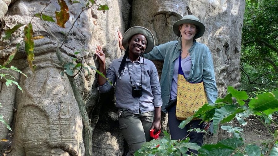 image shows two scientists stand at the base of a large tree, smiling. One is Black and one is European American. They are wearing field gear, hats, and binoculars