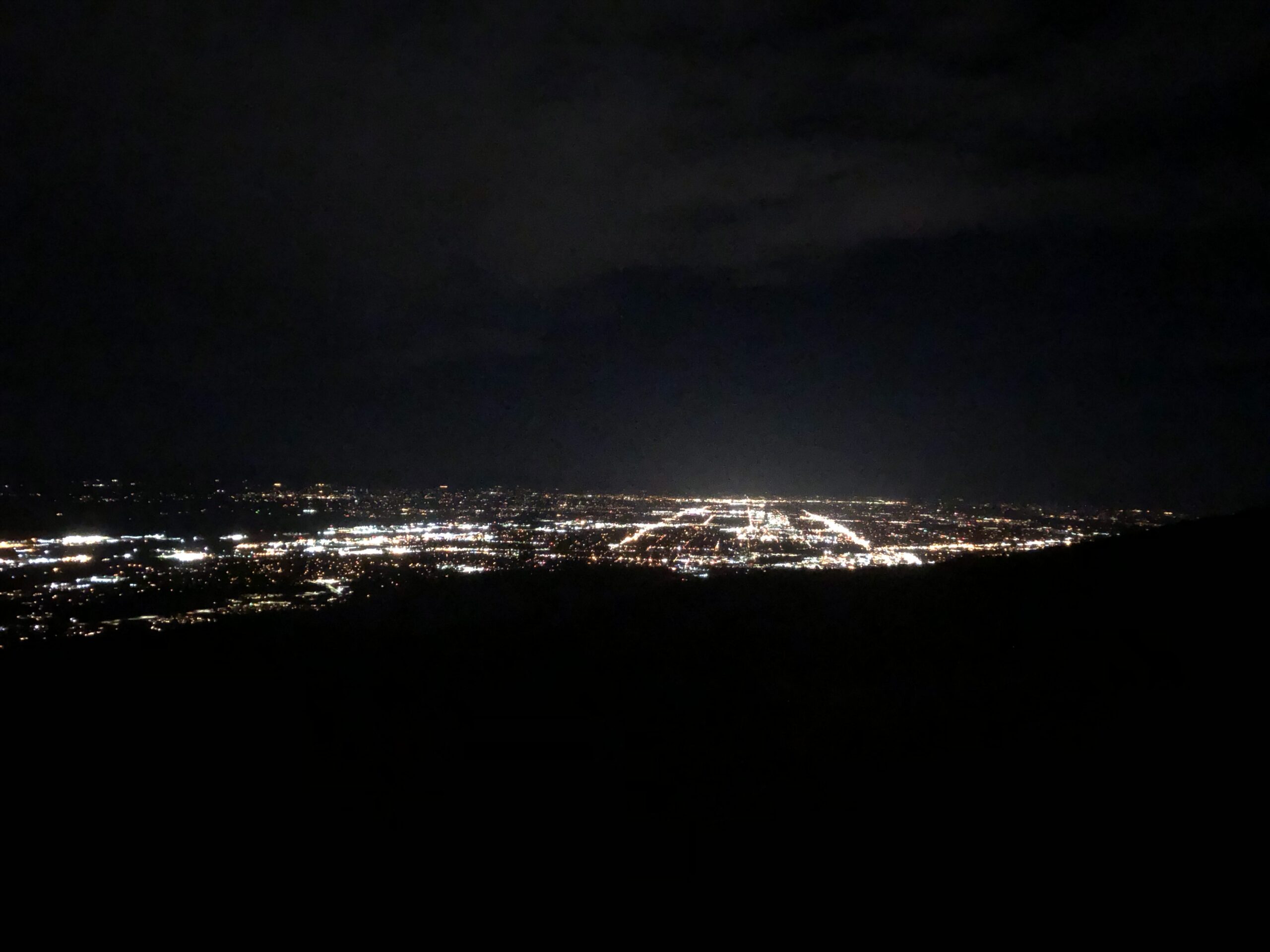 an all dark nighttime image shows glowing city lights in the distance, with the lines from major streets and busy areas shining especially brightly.