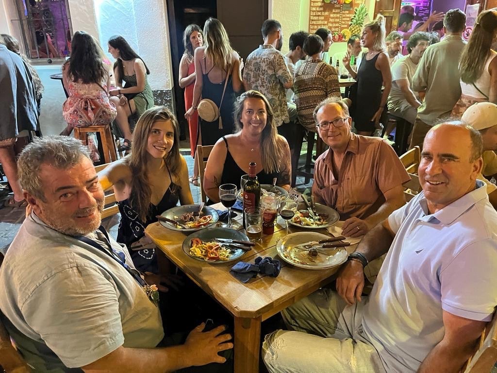 five people sit around a table, smiling at the camera. vibrantly colored food and a bottle of wine rest on the table. In the background is a crowded scene with people chatting and socializing around tables