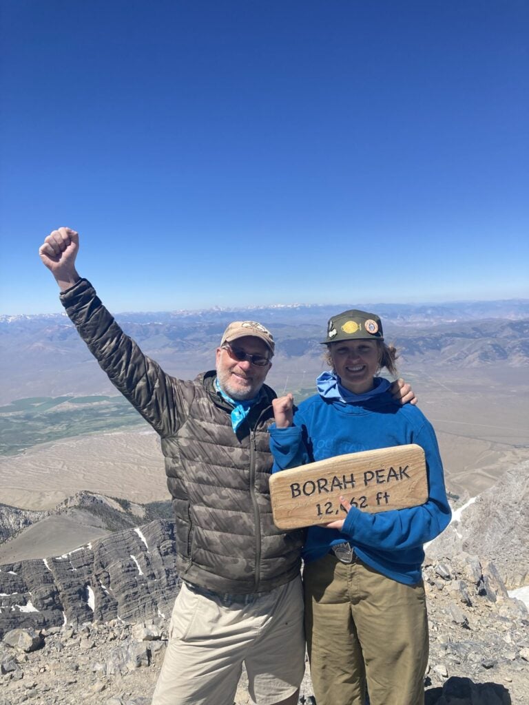 Greg stands with arm raised in triumph next to Ayla who is holding a sign that reads "borah peak. 12662 feet". Behind them the mountains surrounding mount borah are dwarfed in comparison