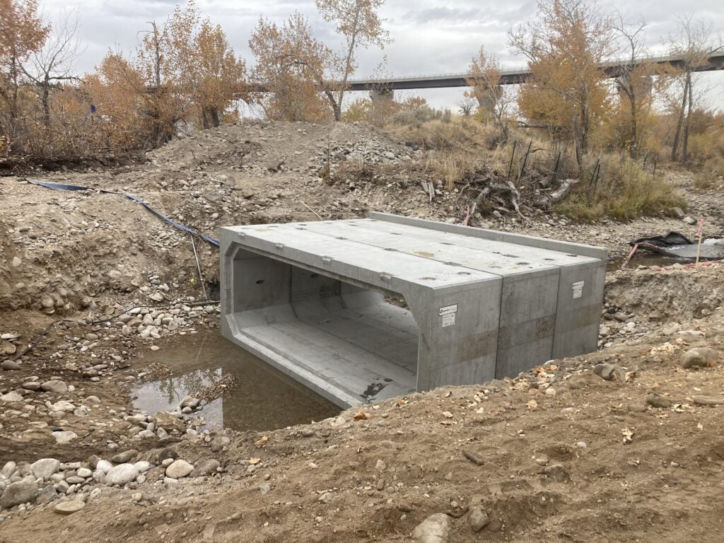 a gray concrete culvert sits down below ground level in an area of excavated dirt. The familiar views of the diane moore nature center including cottonwood trees and the highway 21 bridge are in the background