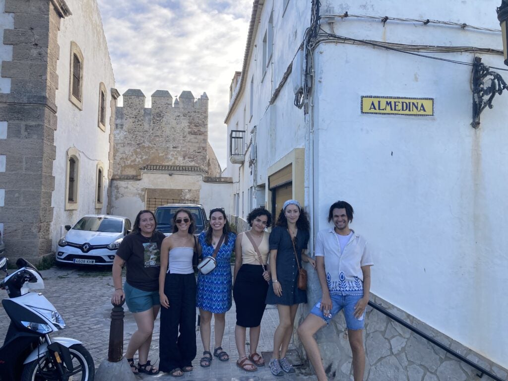 A group of six students standing together smiling. Around them is a classic Spanish street view with cobblestone streets, Wrought Iron balconies, stone buildings, and a decorative tile street sign on a building that reads "Almedina". Small cars and scooters are parked along the narrow street.