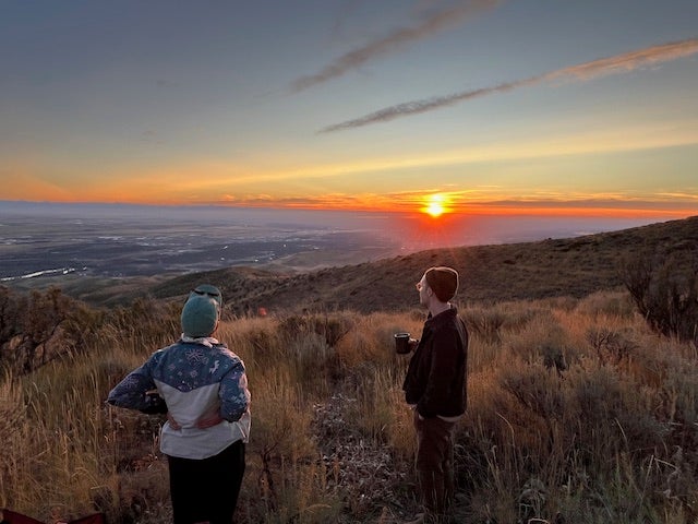 two people stand looking toward a brilliant setting sun. The familiar setting of Lucky Peak's rolling sagebrush hills surrounds them