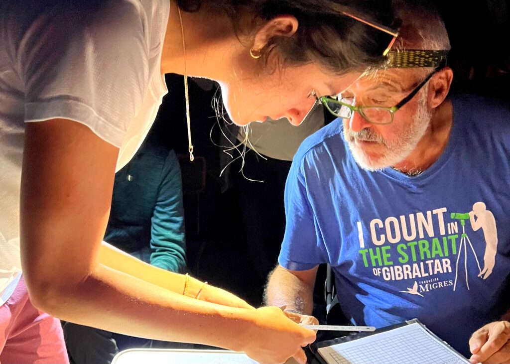 two scientists lean over datasheets. Julia is an undergraduate with long dark hair and small hoop earrings. Alex is an older biologist with gray beard and reading glasses. it is dark around them and they're wearing headlamps. One of the scientist's shirts says "I count in the strait of gibraltar fundacion migres" and has a graphic of a person looking through a spotting scope
