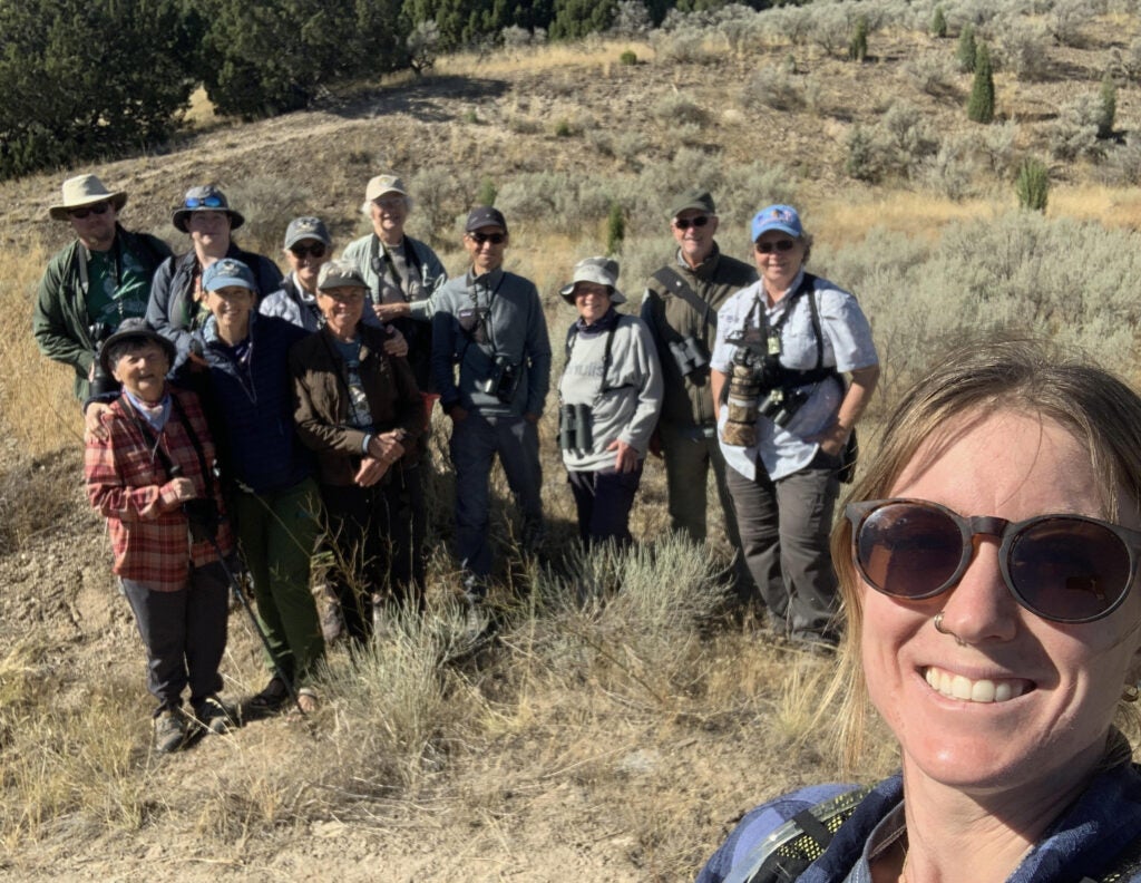 a dozen birders dressed in hats, sunglasses and other field gear stand happily in a group. Lesley, who is taking this photo as a selfie, is in the foreground smiling. Behind the group are some juniper trees and sagebrush.