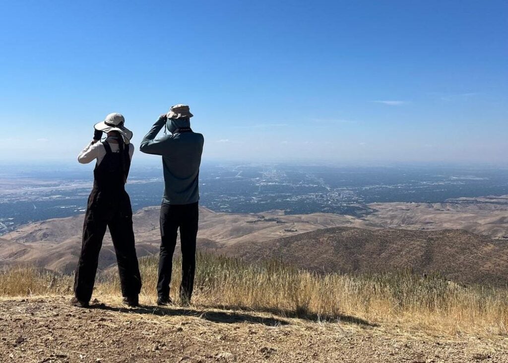 two people stand next to each other facing away from the camera with binoculars raised. They are looking out over Boise which is visible in the background. Both are wearing sun hats and long-sleeved sunshirts beneath the bright sun. The sky is a bright, cloudless blue.
