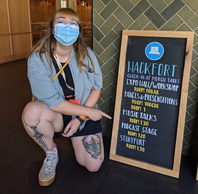Photo of Alisha Graefe kneeling next to, and pointing, to a 'Hackfort' chalkboard.
