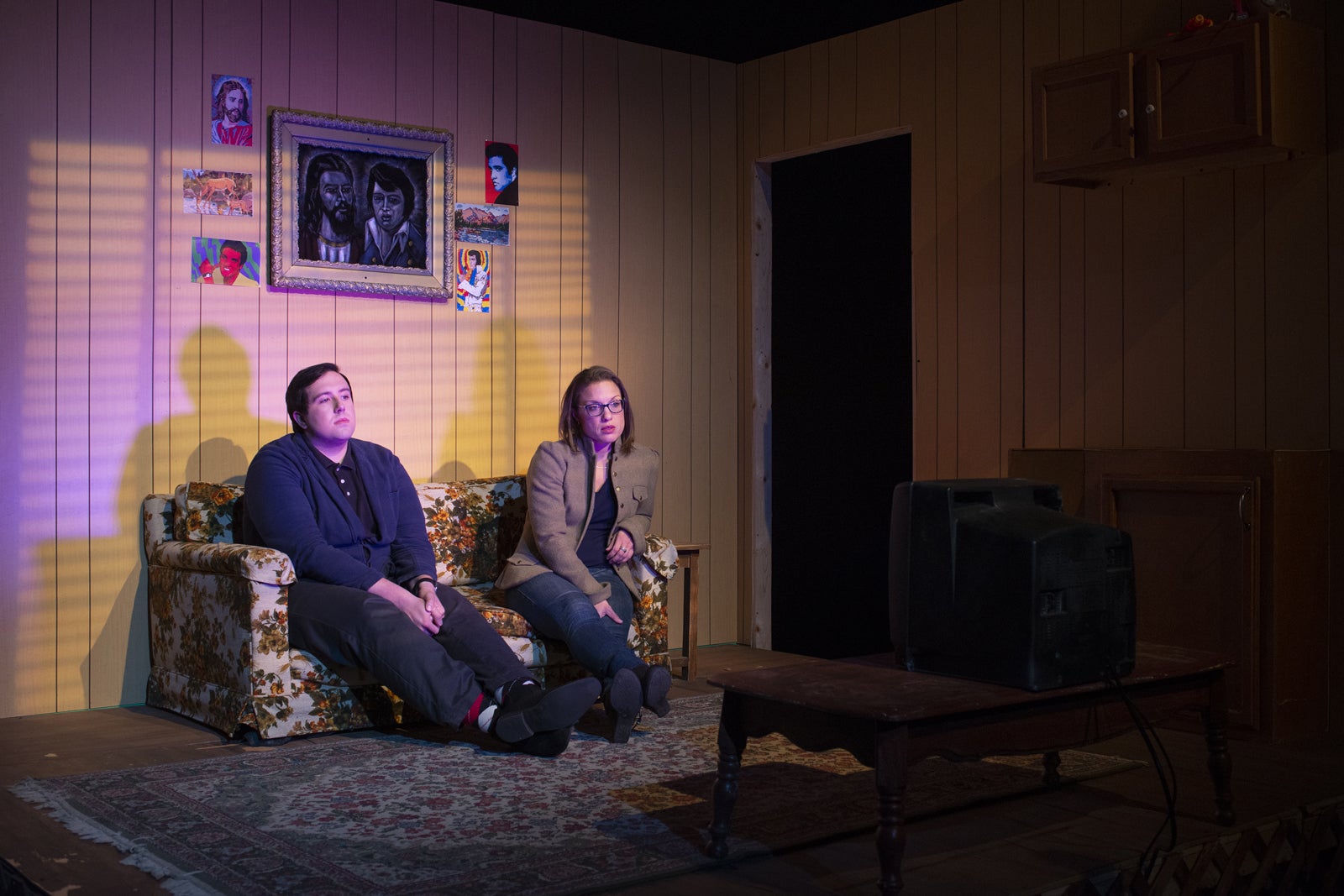 Two actors on stage sitting on a couch staring at TV