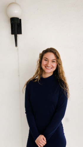 Sandra stands in front of a while wall and smiles at the camera. She is wearing a black long sleeve shirt, and her long, blond curly hair is worn down around her shoulders.