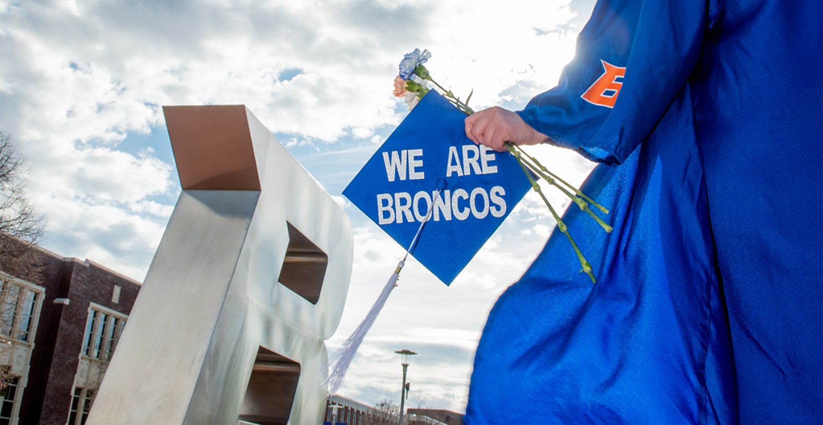 Graduate standing in front of B statue holding flowers and mortar board cap that says We Are Broncos