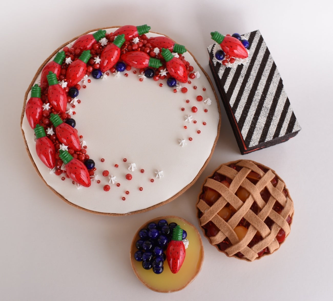 Four desserts arranged on a white surface. There's a white iced round cake with colorful decorations, a black-and-white striped rectangular dessert, a lattice-topped tart, and a yellow-filled round dessert. Each dessert features intricate designs and appealing garnishes.