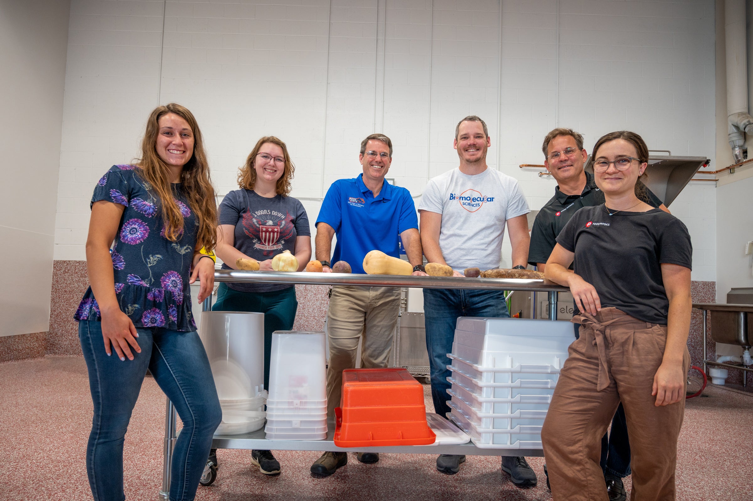 Group photo of six people standing with a table topped with various foods. 
