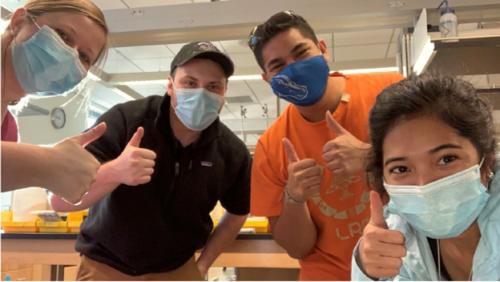 Four people wearing masks give a thumbs up to the camera. 
