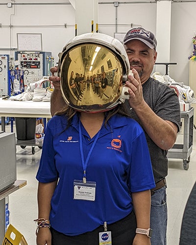 student in blue shirt wears gold screened astronaut helmet, man behinds her helps her put it on